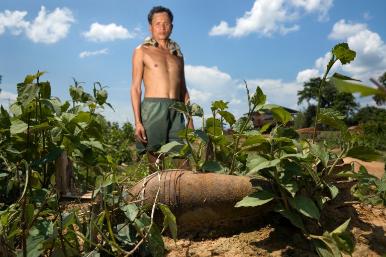 A farmer stands behind a live mortar he found in the field behind his home in Laos, near a branch of the former Ho Chi Minh Trail.