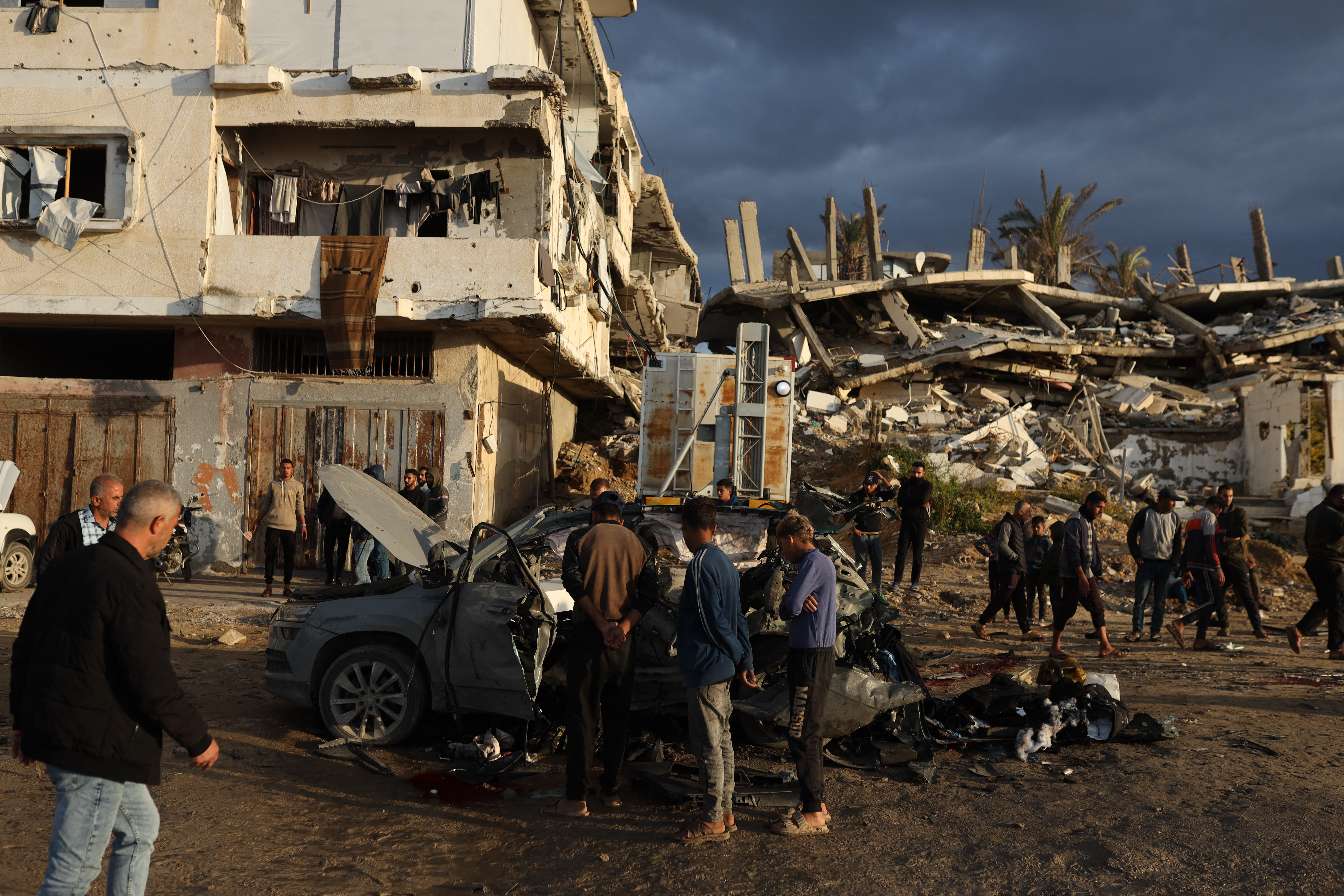 People gather around a destroyed vehicle and rubble after an Israeli airstrike on Al-Rashid Street in Gaza City, Gaza, on December 13, 2025. Local sources and Gaza's civil defense agency reported that four Palestinians were killed in the strike, which occurred during a US-brokered ceasefire between Israel and Hamas that has been in place since October 2025. (Photo by Abood Abusalama / Middle East Images / AFP via Getty Images)